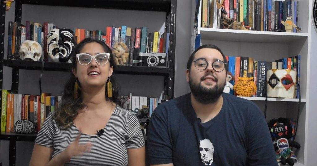 Verônica Domingues e Bruno Motta Mello sentados na frente de uma estante cheia de livros e objetos de decoração